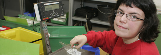 Das Foto zeigt eine junge Frau, die Plastikkleinteile für Schrankscharniere abwiegt. So kann sie die passende Menge anschließend eintüten.