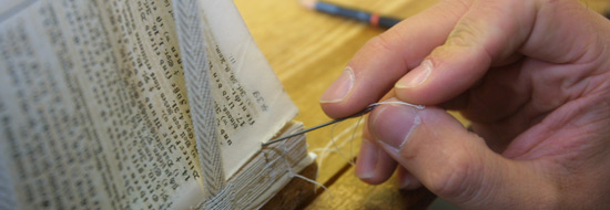 Auf dem Foto ist in Nahaufnahme eine Hand zu sehen, die eine Nadel mit Faden hält. Neben ihr befindet sich in einer Vorrichtung ein kleiner Stapel mit losen Seiten, die zu einem Buch gebunden werden.