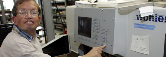 Das Foto zeigt einen Mann, der an einer Papierschneide-Maschine steht und sie programmiert. Vor ihm liegt ein hoher Stapel Papier, der auf das gewünschte Format geschnitten werden soll.