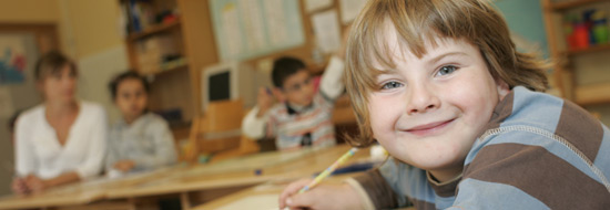 Das Foto zeigt einen kleinen Jungen in seinem Klassenzimmer, der etwas schreibt und dabei ins Bild lächelt.